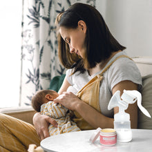 Load image into Gallery viewer, A new mom sits and breastfeeds her new baby with the Organic Nipple Balm and a breast pump next to her on the table.