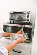 Load image into Gallery viewer, Woman with one hand on microwave and holding hot/cold pack in her other hand.  She is about to microwave the pack.
